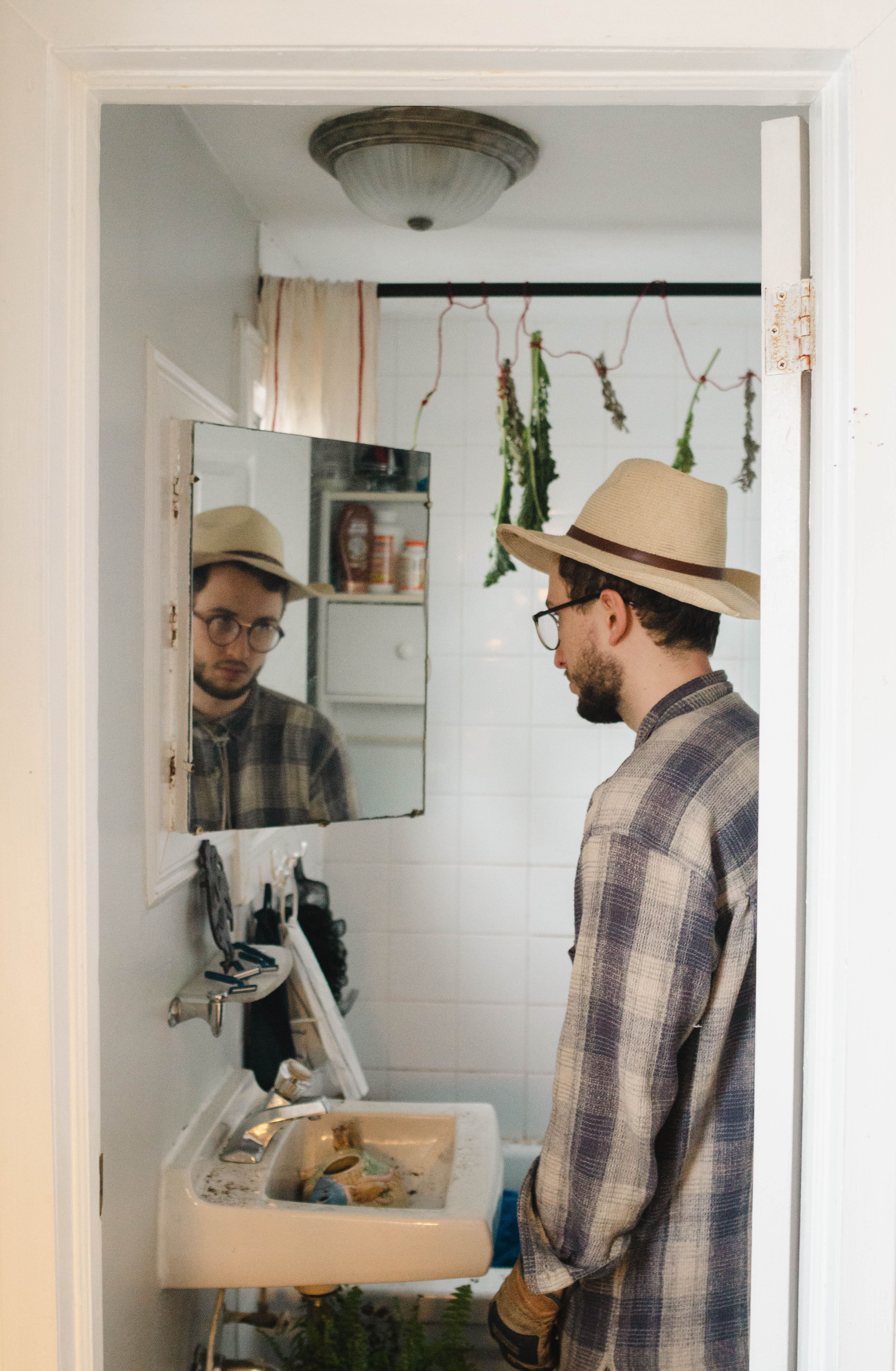 The farmer looks himself in the bathrom mirror. In the background, herbs hang from the shower curtain rod to dry